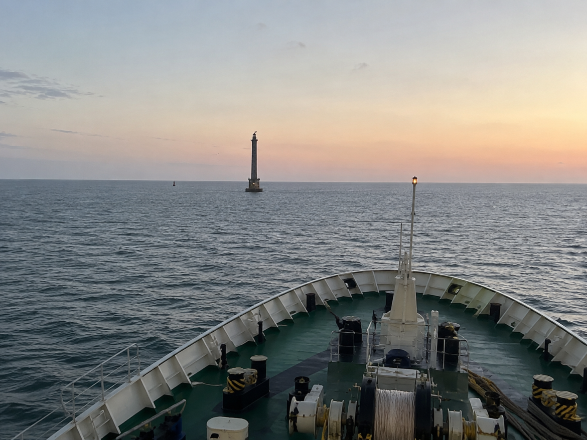 Вид з палуби судна на Аджигольський маяк посеред спокійного Чорного моря на тлі пастельного заходу сонця із зеленою палубою та носовим обладнанням на передньому плані